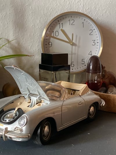 The image depicts a close-up, eye-level shot of a silver toy convertible car with its hood open, revealing a spare tire and engine components. The car is positioned on a dark, flat surface. Behind the car, a round analog clock with a gold bezel is visible, displaying the time as approximately 10:50. To the left of the clock is a black and clear glass bottle labeled "CHIC FOR MEN." To the right of the clock is a small, dark-capped bottle containing a red liquid. The background is a textured, off-white wall. A thin green leaf is partially visible in the upper left corner. The overall impression is a still-life arrangement on a shelf or table, devoid of any human presence or discernible city context. There is no information in the image to suggest a location in Lütjenwestedt, Germany.