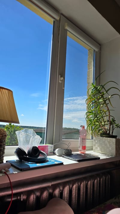 This is a close-up, eye-level photograph taken from inside a room, looking out of a white-framed window onto a bright blue sky with scattered white clouds. The window is divided into two panes by a vertical mullion. The interior of the room features a white windowsill filled with various objects.
On the left side of the windowsill, there is a lampshade, a decorative glass object, and a pair of black headphones resting on a blue and gold patterned tablet case. To the right of the headphones, there is a pink and white box labeled "HEY CLAY". Further to the right, a white electrical adapter is plugged into a white circular device. A tablet lies flat on the windowsill in front of the adapter. Behind the tablet, a clear plastic bottle with a pink label containing a pink liquid and the word "VITAMIN" is visible. To the right of the bottle, a potted aloe vera plant with spiky green leaves sits in a white woven pot. A smaller succulent plant is also in the same pot. The windowsill itself appears to be white.
The background outside the window shows a horizon line with green trees and some buildings, suggesting a rural or suburban setting. Below the windowsill, a dark gray radiator is visible. The bottom right of the image shows a person's leg and foot.
The scene appears to be in Atkočiai, Lithuania, as indicated by the user's prompt. The lighting suggests it is daytime, with bright sunlight casting shadows.