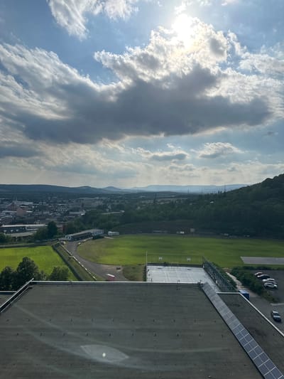This is a high-angle aerial photograph captured in Ústí nad Labem, Czechia, looking out over a sports complex and surrounding urban landscape under a partly cloudy sky with the sun prominently shining through. In the foreground, the dark, flat roof of a building is visible, fitted with rows of solar panels. Adjacent to the roof is a multi-purpose sports court with visible court lines and basketball hoops. Beyond the sports court, a large, green rectangular field, likely a football pitch, is situated. A paved pathway winds around the field, and a few portable toilets are positioned near it. In the middle ground, the town of Ústí nad Labem unfolds, characterized by numerous buildings of varying sizes and architectural styles, interspersed with trees and greenery. The background features rolling hills and mountains, suggesting the city's geographical setting. Several power lines and pylons are scattered throughout the landscape. A parking lot with several cars is visible near the edge of the sports complex. The sky is dynamic, with a mix of bright sunlight and scattered clouds, some of which are dark and heavy, indicating potential weather changes. No people are directly visible in the frame.