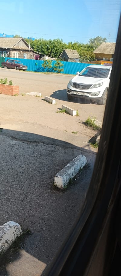 The image depicts an outdoor scene in Киясово, Russia, on a sunny day. In the foreground, a weathered concrete parking bumper is visible on an asphalt surface. Further back, several similar concrete bumpers line what appears to be a parking area. A white Kia Sportage SUV is parked, with its license plate clearly visible as 'Y 404 OA 18'. To the left, a red sedan is parked near a short brick planter. Behind these vehicles, a bright blue metal fence separates the parking area from a residential area. The fence encloses a row of wooden houses with traditional pitched roofs, surrounded by green trees. The sky is a clear, bright blue. The overall impression is of a quiet, sunny day in a rural or semi-rural part of a Russian town.