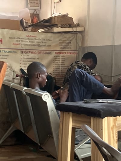 The image shows a partially obscured interior, likely of a small business or training center, located in FUT Minna, Bosso Niger State, Nigeria. Two individuals are visible, partially obscured by furniture. One individual is wearing a patterned shirt and a dark blue jacket and is seated behind the other. A poster behind them advertises computer training, graphic design services (including ID cards, complimentary cards, book publishing, letterheads, and logos), and internet services. The poster lists the address as "Front of Bosso Campus, Beside IBK's Place," and an email: chipsetinfotech@gmail.com. The overall environment appears cluttered and somewhat cramped.