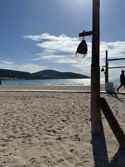 This media file captures a serene beach scene, likely in Anatoliki Attiki, Greece. The image is composed with a strong foreground featuring two prominent wooden posts. The primary vertical post on the left has a woven, basket-like object with brushy fringes hanging from it, suspended by a rope. This post is also wrapped with thick rope near its base. Another angled wooden beam extends from the right side, partially wrapped in rope, creating a rustic, natural aesthetic. The ground in the foreground and mid-ground is a sandy beach, textured with numerous footprints and small undulations.

In the mid-ground, a person is visible walking towards the right, silhouetted against the bright reflection of the sun on the water. A small white crate or low bench is also present on the sand near this individual. Beyond the sand, the shimmering blue sea stretches horizontally, reflecting the bright sunlight. In the background, dark, rolling mountains line the coast, creating a dramatic natural barrier. The sky above is a clear, bright blue, adorned with sparse, elongated white clouds, suggesting a sunny day.

The time of day appears to be daytime, possibly late morning or early afternoon, indicated by the strong sunlight and the reflections on the water. The overall weather is clear and sunny. There is no visible text in the image. The scene evokes a sense of peace and natural beauty typical of a coastal landscape.