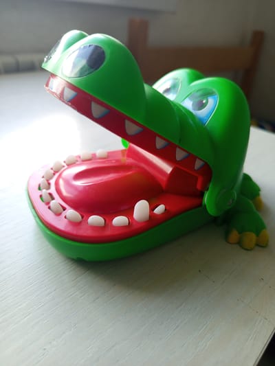 A bright green toy alligator with its mouth wide open is placed on a light-colored wooden table. The alligator's mouth is red inside, with rows of white teeth along the upper and lower jaws. Its eyes are large and cartoonish, with blue irises and black pupils. The alligator has four yellow feet visible, and its body is a vibrant green. The background is out of focus, showing a hint of sunlight coming from a window on the left and the silhouette of a wooden chair on the right. The scene appears to be indoors, on a flat surface, likely a table or desk. The lighting suggests it is daytime. There is no indication of the location being in the city of Кенжекөл, Kazakhstan.