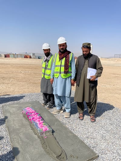 This image, taken in Pir Dust, Afghanistan, depicts three men standing outdoors in what appears to be a construction or industrial site under a clear, bright blue sky.
In the foreground, a rectangular concrete slab rests on a bed of light grey gravel. Embedded within a wet, grey cement strip along the center of this slab is a narrow, metallic, foil-wrapped object adorned with numerous small, bright pink artificial flowers or petals.
Standing behind this slab are three adult men. Two men on the left wear white hard hats and neon yellow safety vests over traditional shalwar kameez. The man furthest left wears a black and white checkered scarf, grey shalwar kameez, and dark sneakers. The middle man wears a red and black patterned scarf, light blue shalwar kameez, and light grey sneakers. The man on the right wears a dark olive green shalwar kameez, a dark vest, and a patterned, round cap, with brown sandals on his feet. He holds a stack of white papers in his left hand. All three men have dark beards and are looking towards the camera. The man in the middle and the man on the right appear to have their adjacent hands close together, possibly clasped.
The background shows a vast, dry, light brown expanse of earth. On the left, a long row of various colored shipping containers (blue, red, brown, white) is visible, some stacked. On the far right, a large industrial structure, possibly a power line tower or a silo, is present. Distant, low-lying hills are visible on the horizon.