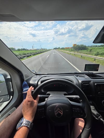 The image depicts the interior of a vehicle on a highway, likely near Kisigmánd, Hungary, given the presence of wind turbines in the distance, a common sight in the region. A person's hands and feet are visible. One hand holds a lit cigarette, and the other rests on the steering wheel, which bears the Citroën logo. A wristwatch is worn on the wrist of the hand holding the cigarette. The feet are clad in striped socks, with one foot resting on the dashboard. The dashboard features a GPS device. The road stretches ahead, with fields and wind turbines visible on either side under a cloudy sky.