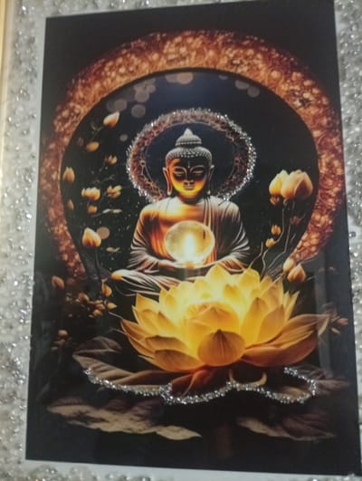 The media file displays a framed artwork featuring a serene Buddha figure seated in meditation. The Buddha is depicted with closed eyes, a gentle expression, and is holding a glowing orb in his hands. He is adorned with a halo of shimmering particles. Surrounding the Buddha is a dark, mystical background with bokeh-like lights. A large, vibrant yellow lotus flower dominates the foreground, with smaller, budding lotuses scattered around the Buddha. The artwork is framed by a decorative border, possibly made of small, reflective elements, giving it a glamorous appearance. The overall mood is peaceful and spiritual. There is no visible text in the image.