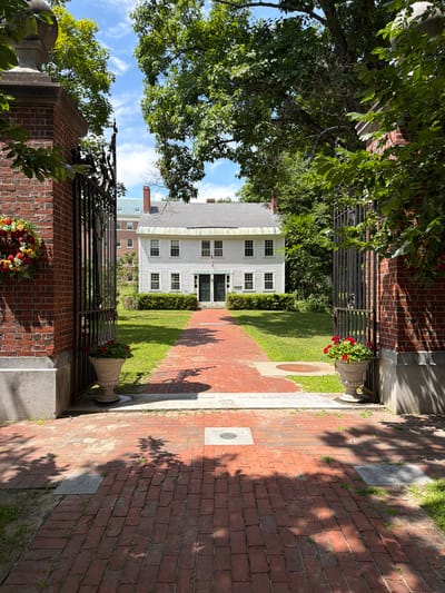 A brick walkway leads through an open wrought-iron gate framed by two brick pillars. A wreath of colorful flowers hangs on the left pillar. Two urns with red flowers sit on either side of the walkway just inside the gate. Beyond the gate, a white, two-story building with multiple windows stands behind a manicured lawn and a hedge. Trees with green foliage surround the building and partially obscure the view. The sky is blue with scattered white clouds. The image appears to be taken on a sunny day.
