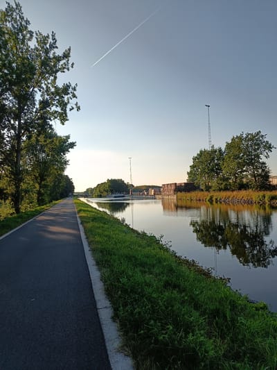 An asphalt path runs along the left side of the frame, bordered by lush green grass and a line of tall trees. To the right of the path is a calm canal reflecting the surrounding trees and structures. In the distance, industrial buildings with stacks of materials are visible, alongside a tall communication tower. A contrail from an airplane stretches across the clear blue sky. The scene appears to be a peaceful riverside path, likely in an industrial area of Haacht, Belgium, during a bright, sunny day.