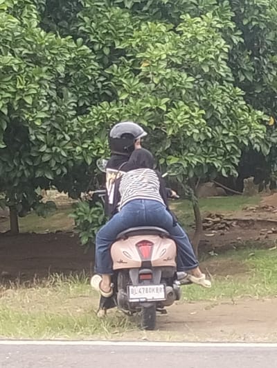 Two individuals are riding a light pink scooter on a paved road in a rural or suburban area, possibly in Teupinreusep, Indonesia. The person in the back, wearing a black and white striped shirt and blue jeans, is seated facing forward. The person in the front, wearing a black jacket and a black helmet, is also seated facing forward. The scooter has a license plate with the characters "BL 4780 KBB." The background consists of lush green trees and foliage, suggesting an outdoor environment. The ground is a mixture of dirt and grass.