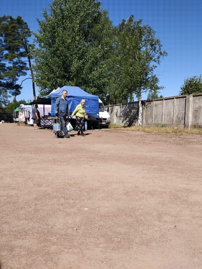 In Irshansk, Ukraine, on a bright, sunny day, a scene unfolds at what appears to be an outdoor market or flea market. The ground is covered in a coarse, sandy soil. In the mid-ground, a blue tent is erected, with a white van parked alongside it. To the left of the tent, another market stall is visible with striped awnings.
Two individuals are walking from left to right in front of the blue tent. The person on the left is a man wearing a striped blue shirt and jeans, pulling a blue and gray shopping cart. The person on the right is a woman in a light green long-sleeved shirt and a black and white patterned skirt, carrying an object in her right hand. Further to the left, a man in a dark t-shirt and shorts stands near another market stall.
The background features a row of large, mature trees, including a tall pine tree on the left and a birch tree with a white trunk. A concrete wall runs along the right side of the image. The sky is a clear, vibrant blue. The overall impression is of a casual outdoor gathering or market activity.