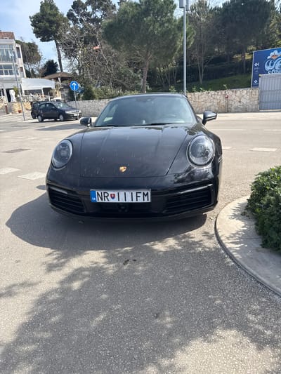 The image shows a black Porsche with the license plate "NR 111FM" parked on an asphalt road. The car is facing the camera and is parked near a curb with greenery behind it. The location is likely in or near the city of Grad Rovinj, Croatia, indicated by the license plate. In the background, there are buildings and trees, suggesting an urban or suburban environment, with a signpost also visible.