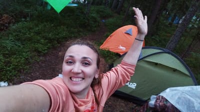 In this medium shot, a woman with curly brown hair and light makeup smiles at the camera while taking a selfie in an outdoor environment. She is wearing an orange-pink long-sleeved shirt. Her right arm is raised, with her hand in a wave. A black bracelet is visible on her wrist. Behind her, an orange tent is set up, and a green tent is partially visible in the foreground. The background consists of trees and dense foliage, suggesting a wooded area. The ground appears to be covered with dirt and leaves. While the image depicts a campsite, specific details placing it in Tarasovskoe, Russia, are not discernible from the visual information alone.