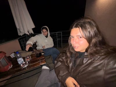 This image depicts a nighttime scene on an outdoor balcony or terrace in Nesso, Italy. Two females are present. In the foreground, a female with long, dark, wavy hair wears a brown leather jacket and is looking directly at the camera, with a metallic object protruding from her lower lip. In the background, a second female, wearing a light-colored hooded sweatshirt and dark jeans, is seated and actively pouring a dark red liquid from a dark bottle into a clear tumbler on a wooden table. A lit cigarette is visible in her mouth. The wooden table holds a red lantern-like object, a small white packet, a clear plastic bottle labeled "FONTARIELLA" (water), and three clear tumblers, two containing liquid and one receiving the poured liquid. An off-white patio umbrella is partially visible in the upper left, and a metal railing is situated behind the table. The background beyond the railing is entirely dark, indicating night. A light-colored wall is visible on the right side of the frame.