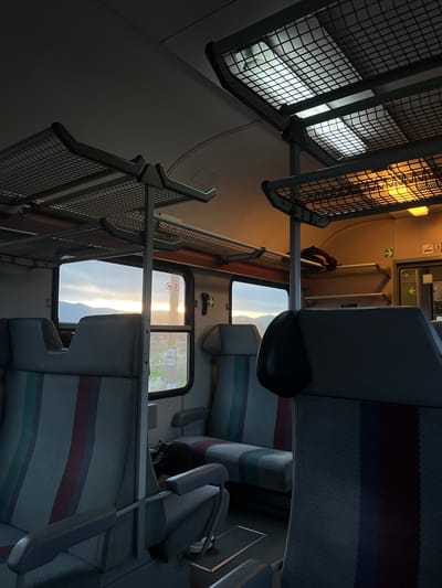 The interior of a train carriage is depicted with rows of seats upholstered in a striped pattern. Overhead luggage racks, constructed from metal frames and mesh, line the upper section of the carriage. The windows offer a glimpse of an exterior scene, possibly mountains illuminated by the setting sun. No people are clearly visible, but a bag is on the seat. The location is unknown; there is no visual evidence to link to Polyanovo, Bulgaria.
