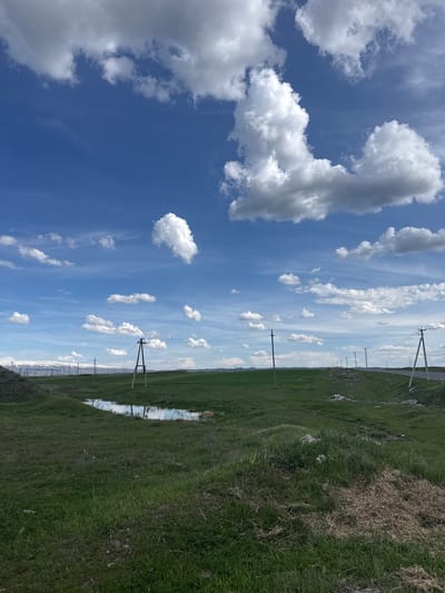 The image depicts a daytime scene in Getk, Armenia, featuring a clear blue sky with scattered cumulus clouds. The ground is covered in green grass, with some patches of dry vegetation. A series of utility poles with power lines stretch into the distance. A narrow body of water, possibly a ditch or small stream, reflects the sky and clouds. No people or animals are visible. The overall environment appears rural or agricultural.