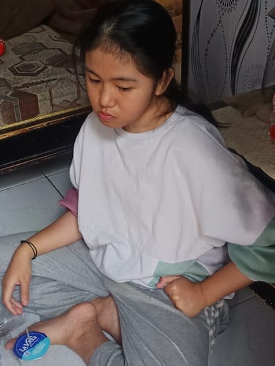 The image displays a young female subject seated on a light-colored tiled floor indoors, located in Limbangan Sari, Indonesia. The female has dark hair, pulled back from her face, and is looking downwards with a neutral expression. She is wearing a light-colored, possibly white, long-sleeved crew-neck shirt over an inner garment with pink and light green sections, paired with grey casual trousers. Her feet are bare, and a black elastic band is present on her left wrist. Her left hand rests on her left leg, adjacent to a small, transparent plastic container with a blue cap labeled "La Vida." Her right hand is clenched into a fist, resting on her right leg. In the background, to the left, a dark wooden furniture item with a patterned cushion (featuring brown, beige, and black geometric shapes) is visible. To the right, a wall covered with white wallpaper displaying a black and white abstract floral or swirl pattern is present. A light-colored woven mat and a small bottle with a red top are partially visible on a surface in the far right background.