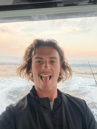 A man poses with his tongue out in an outdoor setting. His hair is blond and wavy, and he's wearing a dark jacket. A railing is visible on the left. The background features a body of water with waves, possibly the ocean, under a sky with soft, muted colors, suggesting it could be sunrise or sunset. A fishing pole is also visible in the background. The man appears to be enjoying himself.