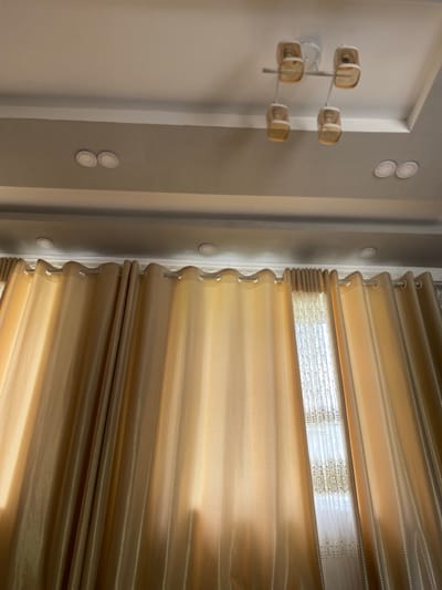 The image captures an interior scene, looking up towards a ceiling and a window draped with curtains. The ceiling is painted white and features recessed lighting fixtures. A modern light fixture with four amber glass shades hangs from the ceiling. The window is covered by two layers of curtains: sheer white lace curtains with decorative trim are layered behind heavier, golden-brown curtains. Sunlight filters through the sheer curtains, casting a warm glow. The curtains are hung from silver rods. The scene is consistent with a residential interior in Kyshtut, Kyrgyzstan.