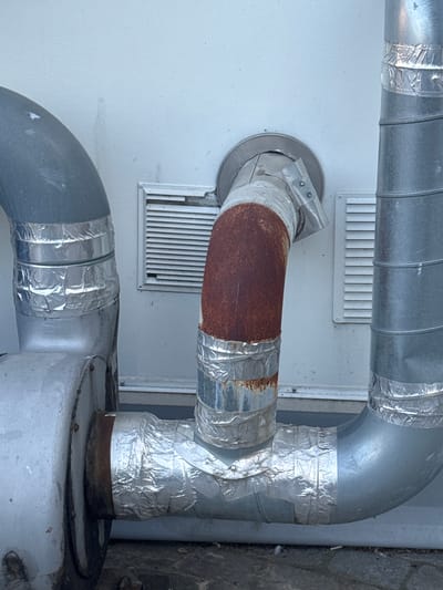 The image shows an exterior view of industrial ventilation pipes. The pipes are made of metal, with some sections showing signs of rust and wear. They are insulated with reflective foil tape in several places. Two rectangular ventilation grilles are visible on a light-colored wall behind the pipes. The scene appears to be located in an urban or industrial setting. The image was taken in Strombeek-Bever, Belgium.