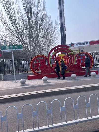 This image captures a daytime urban scene on Lingdong Jie in Huanggu District, China, under a clear, bright blue sky. The foreground features a light gray metal pedestrian barrier with arch-top sections. Immediately behind this is an asphalt roadway, bordered by a paved sidewalk constructed of light gray and darker gray sections. Four spherical, light gray stone bollards are spaced along the sidewalk's edge.

On the sidewalk, a large, ornate red and gold decorative structure stands prominently. It is composed of circular frames containing stylized Chinese characters (identified as "福" and "春"), fan motifs, and a central lantern image. To the left of this display, a green street sign clearly labels the location as "陵东街 (LINGDONG JIE)", indicating both Chinese and Pinyin transcription.

Three individuals, all wearing face masks, are present on the sidewalk. Two people, dressed in dark winter-style clothing, walk from right to left (westbound) past the decoration. One carries a dark shoulder bag. A third person, wearing a dark jacket and lighter pants, walks from left to right (eastbound) further to the right of the decoration.

Behind the sidewalk and decorative elements, several tall, bare deciduous trees are visible, indicating a winter or early spring season. In the background, to the right, a "中国融通" (China Rongtong) service station with a red star logo is visible, alongside a tall, multi-panel advertising pylon.