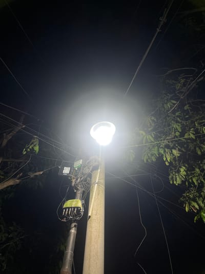The image shows a brightly lit street lamp in Binong, Indonesia. The light fixture is mounted on a utility pole with numerous wires and electrical boxes attached. Surrounding the pole are trees and other vegetation, suggesting a residential or semi-rural area. The time of day is night.