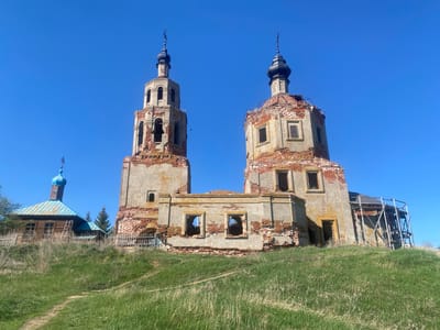 The image shows a partially ruined brick and concrete church in Шуран, Russia. The church has two prominent towers with onion-shaped domes topped with crosses. The structure is heavily weathered and damaged, with exposed brick and missing sections of the outer walls. There are multiple window openings without glass or frames. In the background, to the left of the church, is a smaller wooden building with a blue dome and a wooden fence surrounding the property. The entire scene is set on a grassy hill, with a worn path leading up to the buildings. The sky is clear and blue.