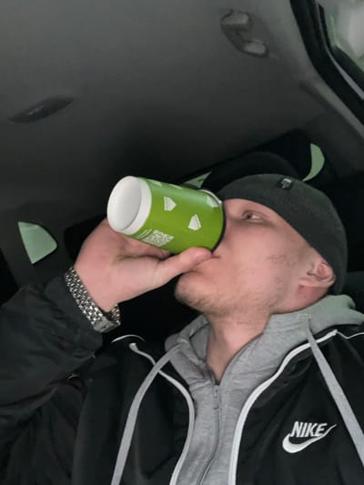 An adult male is observed inside the cabin of a vehicle, positioned in Dobryanka, Russia. He is depicted in a side profile, looking upwards while actively drinking from a bright green disposable paper cup. The cup features white geometric shapes and a visible QR code. The male is wearing a dark grey beanie, a black athletic jacket with a white Nike logo on the right chest, and a grey hooded sweatshirt underneath. A silver-colored metal link watch is present on his left wrist. The vehicle's interior headliner, dark grey in color, is visible above him, along with overhead fixtures. The background is dark, indicating low light conditions within the vehicle.