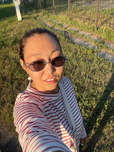 In an outdoor setting, a woman of East Asian descent takes a selfie. She has her hair pulled back and is wearing sunglasses and a white and red striped long-sleeved shirt. A light-colored strap from a bag is visible across her shoulder. She is standing on a path with grass and some gravel. In the background, there is a fence and some buildings. The lighting suggests it is late afternoon or early evening. The location is identified as Кішкенекөл ауылдық округі, Kazakhstan.