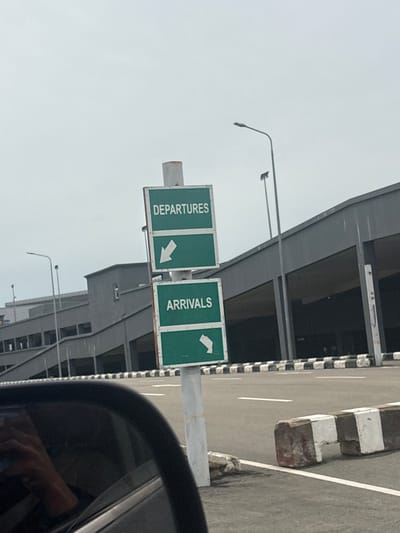 In Gwaska, Nigeria, an airport signage directs traffic. The signs are green with white text and arrows, indicating "DEPARTURES" with an arrow pointing left, and "ARRIVALS" with an arrow pointing down-left. The signs are mounted on a white pole. In the background, a modern airport terminal building with large, dark openings is visible. The sky is overcast. A street runs in front of the terminal, marked with white lines and a black and white checkered barrier. Part of a car's black interior, including a reflection of a hand, is visible in the foreground.