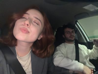Two individuals are visible inside a vehicle. A male is wearing a light-colored long-sleeved shirt with a black seatbelt across his chest. A female with red hair is next to him. The interior of the vehicle and a portion of the windshield are visible. The location is unknown.