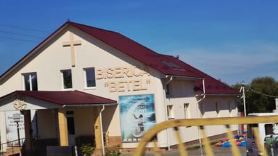 The image captures a church building with a burgundy roof and light-colored walls, prominently displaying the words "BISERICA BETEL" in large, tan letters. A large cross is mounted on the gable. Below the text, a large poster is affixed to the wall, featuring the words "NOU ÎNCEPUT" (New Beginning) in a dramatic blue and white design, with a silhouetted figure reaching upwards. To the left of the main entrance, another poster or sign is partially visible, with text in Romanian. The building has several windows and a covered entrance. In front of the building, a yellow railing is visible, partially obscuring a section of the scene. Beyond the railing, children's toys in bright colors, such as red, blue, and yellow, are scattered on the ground, and a white van is parked. The background shows a clear blue sky with some wispy clouds and a cluster of trees. This is the Bethel Church located in Ialoveni, Moldova.