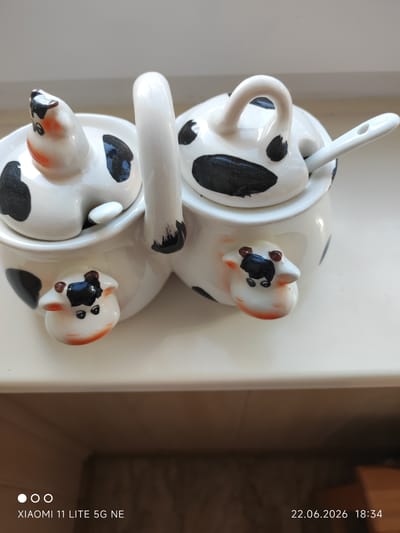 This is a close-up, eye-level shot of a novelty ceramic sugar and creamer set designed to look like cows. The set is white with black cow spots and features small, molded cow faces on the front of each container. The creamer, on the left, has a cow's head lid with orange cheeks and black eyes and snout. The sugar bowl, on the right, also has a cow's head lid and is accompanied by a small white spoon that rests in a hole in the lid. A white ceramic handle connects the two containers. The set sits on a light-colored surface, possibly a windowsill, with a blurred background of a wall and a darker area below. The lighting suggests it is daytime. The bottom right corner of the image displays the text "22.06.2026 18:34" and the bottom left corner shows "XIAOMI 11 LITE 5G NE".