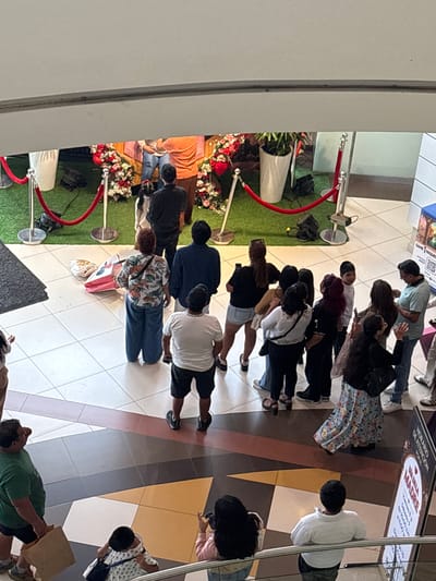 The image shows a group of people queuing in what appears to be an indoor space, possibly a shopping mall or event center. They seem to be observing or waiting for something on the left, which is partially obscured by a red rope barrier and decorations, possibly a floral display. There are potted plants and what seem to be artificial grass sections. Some individuals are looking at their phones. The ground has a tiled pattern. A sign related to "Dia de la Madre" (Mother's Day) is visible in the upper right. Based on the sign referencing Spanish and the location details provided, this scene likely takes place in a commercial area in San Juan de Miraflores, Peru.