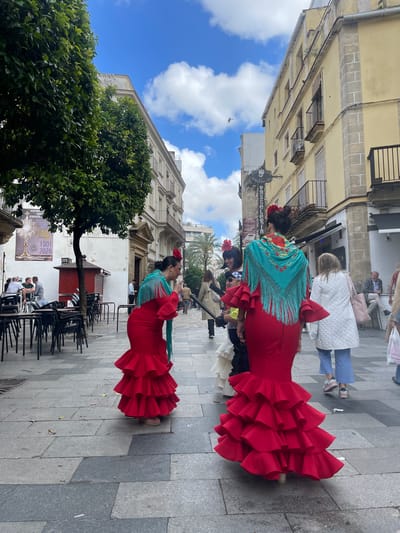 The image captures a street scene in Jerez De La Frontera, Spain. Two women are prominently featured, wearing vibrant red flamenco dresses with ruffled skirts and turquoise shawls adorned with floral embroidery. Both have their hair up with a red flower. They are standing on a paved street with a backdrop of buildings with a bright blue sky and white clouds above. A large green tree is to the left of the lower figure. The street appears to be a pedestrian zone with people walking in the background, and there are tables and chairs of an outdoor cafe in the foreground.
