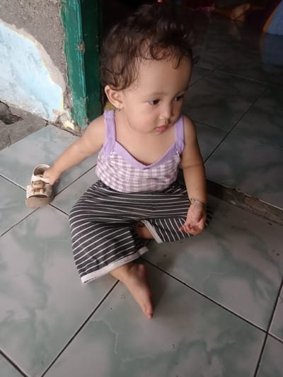 A young child is sitting cross-legged on a tiled floor in what appears to be a residential area. The child has curly hair and is wearing a light purple and white checkered tank top with grey and white striped pants. The child is holding a small sandal in their left hand. The background shows a wall with peeling paint, suggesting an older building. Based on the location provided, the scene is in Bungur, Indonesia.