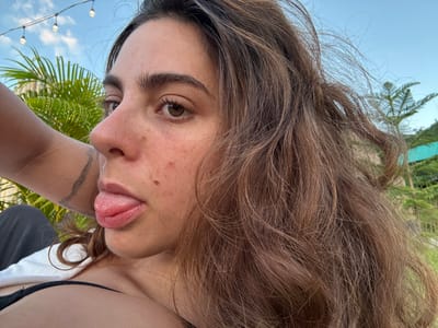 The image presents a close-up, eye-level perspective of a female individual outdoors in Ban Mae Ping, Thailand. The subject has long, wavy brown hair, brown eyes, and a fair complexion, exhibiting visible moles, freckles, and minor skin blemishes on her left cheek. Her head is slightly tilted, and she is looking forward with her tongue extended. She is wearing a dark-colored top, with a strap visible over her left shoulder. To the immediate left, a portion of another individual's left arm is visible, tattooed with a curvilinear design. The background shows a clear blue sky above lush green palm trees and other foliage. String lights are horizontally strung across the upper left background. A light-colored piece of fabric is visible in the lower left foreground.