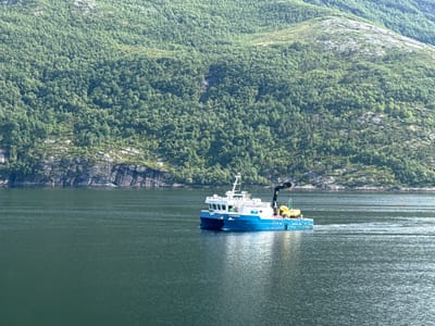 An aerial view captures a bright blue and white vessel, identified as "STERKEFIK" by the lettering on its hull, navigating through a dark, choppy body of water. The boat is equipped with a crane and what appears to be yellow buoys or flotation devices. In the background, a steep, lushly forested mountain slopes down towards the water's edge. The mountain is characterized by its dense green foliage interspersed with rocky outcrops and lighter green patches, suggesting varied terrain. The sky is not visible in the frame. The scene suggests a location within a Norwegian fjord, given the characteristic mountainous landscape meeting the water.