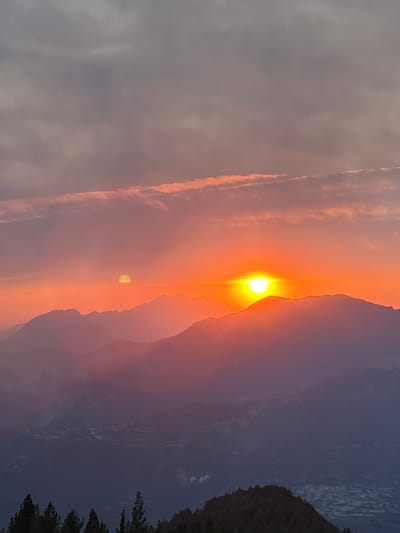 A scenic sunset unfolds over a mountainous landscape, with layers of hazy mountains receding into the distance. The sky is a vibrant gradient of orange and pink, transitioning to gray at the top, with wispy clouds. Two distinct suns are visible: a bright, intense sun partially obscured by a mountain ridge on the right, and a fainter, yellow orb higher and to the left. In the foreground, a dark silhouette of trees lines the bottom of the frame, with the faint outlines of a town or village visible in the far distance below the mountains. This scene is located in the vicinity of San Juan de Plan, a village in the Spanish Pyrenees, where the mountains provide a dramatic backdrop for the sunset.