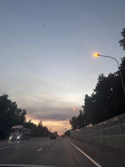 The image shows a highway with several vehicles on it, including a large truck on the left and a car in the distance on the right. The sky is a gradient of pale blue to orange, indicating either dawn or dusk. There are trees lining the side of the road, and streetlights are visible. A sound barrier is present along the right side of the highway. The scene appears to be set in a semi-urban or suburban area.