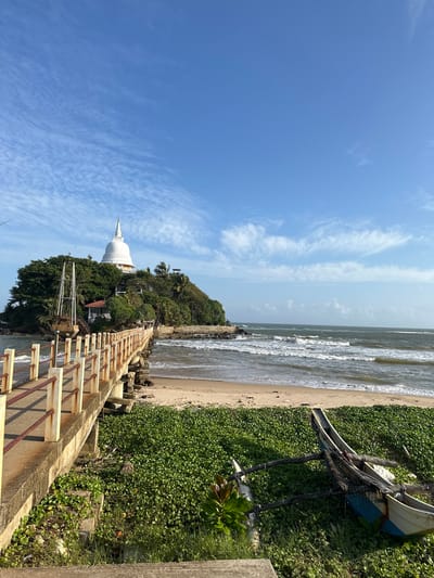 This image depicts the Buddhist temple on the small island of Paravi Duwa, located in the city of Matara, Sri Lanka. A concrete bridge with weathered red railings connects the mainland to the island. The island is densely covered with lush green trees, and a prominent white stupa crowns its summit. To the left of the stupa, a weathered structure resembling a ship's mast is visible. Below the bridge, the sandy beach meets the ocean, with moderate waves breaking near the shore. In the foreground, on the right side of the image, a traditional outrigger canoe rests on a carpet of green vegetation. The sky above is a clear blue with scattered wispy clouds.