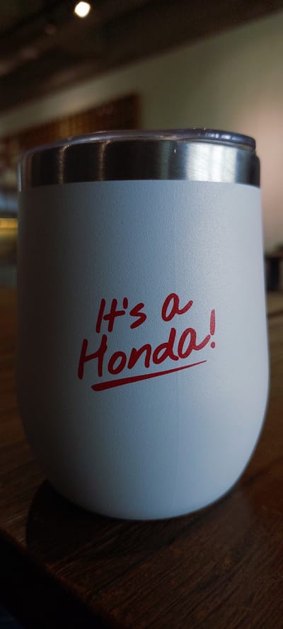 The image shows a white tumbler with red lettering that reads "It's a Honda!". The tumbler has a silver rim and is placed on a dark wooden surface, possibly a table. The background is out of focus, showing hints of a ceiling with lights. There are no people or other discernible objects in the image. The lighting suggests it is indoors, possibly in a cafe or restaurant. The image does not provide any information or context related to Cicalengka, Indonesia.