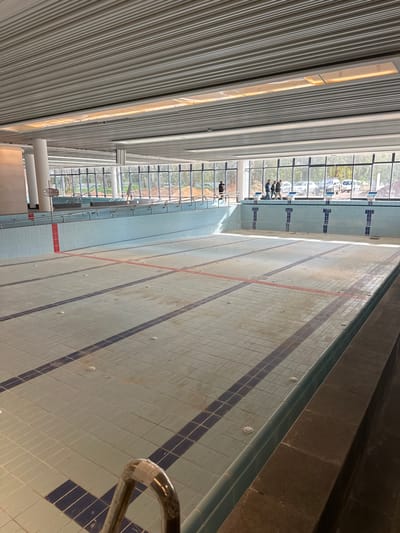 An indoor swimming pool facility in Kadima-Zoran, Israel, is depicted with a large, rectangular main pool emptied of water. The pool floor is tiled in light blue and white, featuring dark blue lane lines and a red line marking the shallow end. Multiple circular fixtures are evenly spaced on the pool floor. A chrome-plated pool ladder is visible in the lower left foreground. Along the far end of the pool, five dark-colored starting blocks are installed. A system of chrome handrails extends along the pool's edge, leading to a shallow-end access ramp on the left, next to which a "No Diving" sign is posted.
The facility features a high, light-colored slatted metal ceiling with integrated linear lighting. Large white cylindrical support columns are visible on the left. The far wall consists of floor-to-ceiling windows, revealing an exterior environment with trees, disturbed earth, and several parked automobiles.
Five individuals are present near the far pool edge. One individual, dressed in dark clothing, stands alone on the left side of the pool deck. A group of four individuals, also dressed in dark or casual attire, stands together further to the right, near the windows, appearing to observe the scene or engage in conversation. No specific actions or interactions beyond standing are discernible. The overall scene suggests a newly constructed, drained, or undergoing maintenance aquatic center.