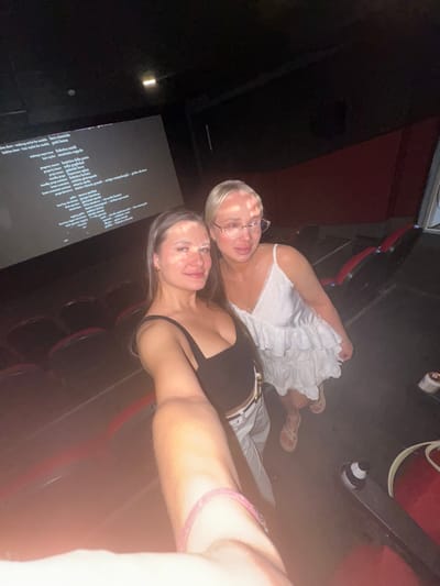 Two women are taking a selfie inside a movie theater. One woman is wearing a white dress and glasses, while the other is wearing a black top. The movie screen is visible in the background, showing the film credits. The location is Lesnoi Gorodok, Russia.