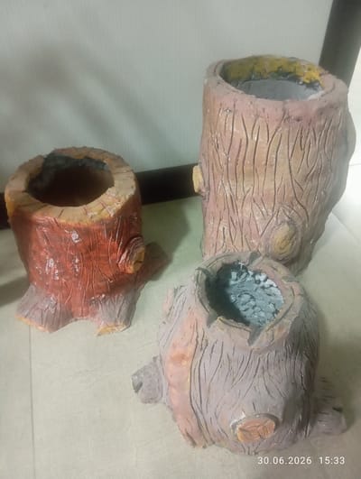 Three decorative pots resembling tree stumps are arranged on a light-colored tiled floor. The pots are made of a material that mimics wood grain and bark. The largest pot, positioned in the center-right of the image, is a light brown with darker brown vertical lines and a prominent knot detail. Its opening is visible, and it appears to contain small gray pebbles. To its left and slightly in front is a smaller pot, predominantly reddish-brown with darker grooves and a knot detail. Its opening shows a dark interior. In the foreground, to the right of the reddish-brown pot, is another small pot, similar in shape and texture to the large central one, with a light brown and gray mottled exterior and an opening filled with gray pebbles. The background consists of a plain, light-colored wall. A dark vertical object, possibly a doorframe or furniture, is visible on the far right of the background. A timestamp "30.06.2026 15:33" is overlaid on the bottom right of the image. The scene is indoors and well-lit. There are no people present in the image. The image does not provide any information about the city of Ohangaron, Uzbekistan.