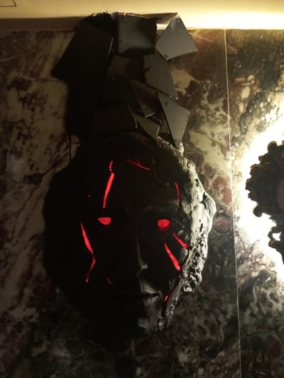 The image displays a dark, stylized, mask-like sculptural wall art piece, approximately 70-80cm in vertical dimension, mounted on a polished marble wall within an interior location in Bashkia Durrës, Albania. The sculpture, rendered in a rough, dark grey or black material, presents a fragmented human face with glowing red elliptical eyes and prominent, irregular red light fissures extending across its surface, resembling cracks or lava flows. Above the face, a cluster of non-uniform, dark, angular planar fragments, some visibly attached with small fasteners, forms a jagged crest. The surrounding wall consists of marble tiles exhibiting mottled white, grey, and reddish-brown veining. A warm, linear light source, positioned above the artwork and just out of frame, illuminates the upper portion of the wall and sculpture. A partially visible, ornate, dark-colored object is present on the right edge of the frame, mounted on the same marble wall. No individuals or dynamic actions are present.