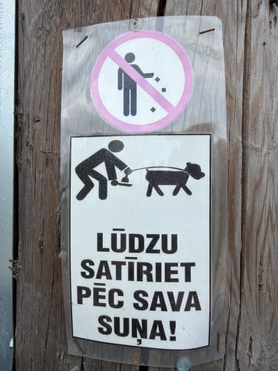 This media file features a sign affixed to a weathered wooden surface, likely outdoors given the texture of the wood and the presence of a plastic covering over the sign. The sign is divided into two distinct sections.
The left section displays a circular prohibition symbol with a pink border and a pink diagonal line. Inside the circle, a stylized human figure is depicted, bent over, with small squares scattered around it, suggesting the act of urinating. This symbol is commonly understood as a "no urination" sign.
The right section of the sign contains a graphic and text. The graphic depicts a silhouette of a person holding a dog on a leash. The person is bent over as if to pick up something, and the dog is standing beside them. The text below this graphic is in Latvian and reads: "LŪDZU SATĪRI PĒC SAVA SUŅA!". This translates to "PLEASE CLEAN UP AFTER YOUR DOG!".
The overall scene conveys a directive regarding sanitation and responsible pet ownership. The wooden background appears to be part of an outdoor structure or fence. There are no specific indicators of time of day or weather conditions, but the aged appearance of the wood suggests it has been exposed to the elements. The location context provided is Tēraudi, Latvia.