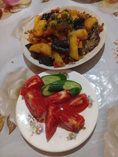 The image displays a meal laid out on a table. In the foreground, a white plate with a floral pattern contains several slices of fresh tomatoes and some cucumber slices. Behind this plate, another white plate holds a dish of cooked vegetables, primarily potatoes and eggplant, seasoned with herbs. The food is arranged on a patterned tablecloth. The setting appears to be a domestic environment, likely a home, and given the context provided, it is situated in the city of Tursunzoda, Tajikistan. No people are visible in the image.