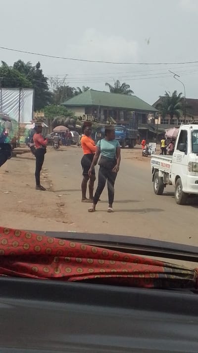 This image depicts a daylight street scene in Igbo Ukwu, Nigeria. The perspective is from inside a vehicle looking out onto a roadside environment.

**People:** Three adult females are prominently positioned in the mid-ground, standing near the unpaved edge of a road. The leftmost female wears an orange long-sleeved shirt and black trousers, carrying an unidentified flat object on her head, and stands with arms crossed, facing right. The central female wears an orange short-sleeved top and black shorts, carrying a woven, light-colored bundle or basket on her head, and stands with arms crossed, facing left, wearing light-colored open-toed sandals. The rightmost female wears a light blue short-sleeved top and black trousers, also carrying a woven, light-colored bundle or basket on her head, with a dark bag slung diagonally across her body. She stands facing slightly left, looking right, and wears light-colored open-toed sandals. Additional individuals are visible in the background, including one person in a yellow vest near a vehicle and others near motorbikes and market stalls.

**Objects:** A white pickup truck, marked "KIZPAREX TABLE WATER" and displaying a partial phone number, is positioned on the right side of the road, facing left. A blue truck is visible in the distant background on the left. Power lines extend horizontally across the upper frame, and a street light pole is present on the right. The bottom of the frame reveals a partial view of a red patterned fabric and a black surface, consistent with a vehicle's interior.

**Environment:** The street features both unpaved and paved sections. Buildings with varying roof styles, including one with a green sloped roof, line the street. Numerous green trees and palm trees are interspersed throughout the background. The sky is overcast and grey.