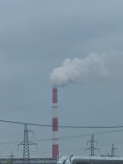 In Luchegorsk, Russia, a towering industrial smokestack, painted with alternating red and white bands, releases a thick plume of white smoke into a hazy, overcast sky. The smokestack is positioned to the right of the frame. In the foreground, a network of electrical power lines and transmission towers crisscrosses the scene, with the base of the smokestack partially obscured by these structures. The overall impression is of an industrial landscape under a muted, gray sky.