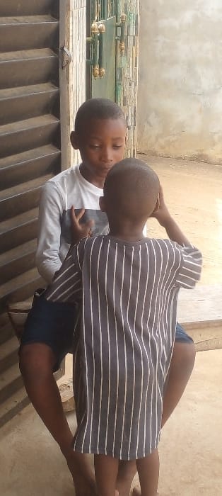 Two young males are depicted in Agunmo, Nigeria. The taller male, wearing a white long-sleeved shirt with a horizontal grey stripe across the chest and dark shorts, is seated on a light-colored concrete ledge. He looks downwards or towards the other child, with his right hand raised in front of his chest, fingers slightly bent. A shorter male stands directly in front of him with his back to the viewer, wearing a dark grey or black short-sleeved shirt with vertical white stripes. The standing child has both hands raised to the sides of his head. Both children appear to be barefoot on a light-colored concrete surface. The background features a partially open brown louvered shutter on the left and a green metal gate with gold-colored decorative finials to the right, set against a light-colored, possibly textured, wall.